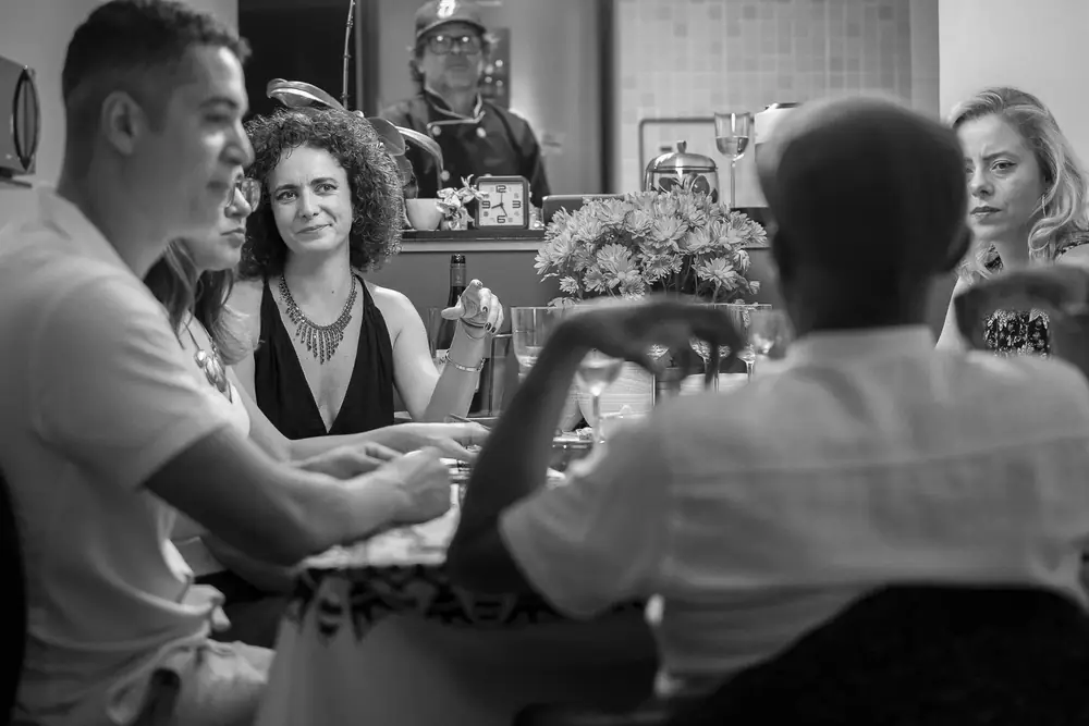 Convidados conversando à mesa, com o chef ao fundo, em preto e branco